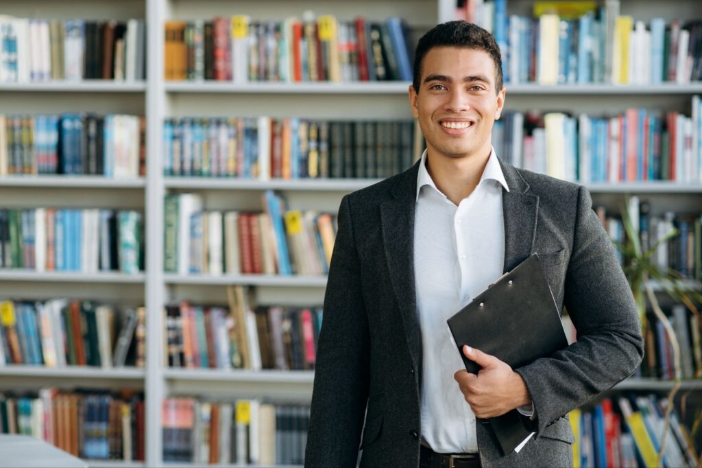A smiling male professional in a gray blazer and white shirt, holding a black clipboard in front of floor-to-ceiling bookshelves in a modern library or office.